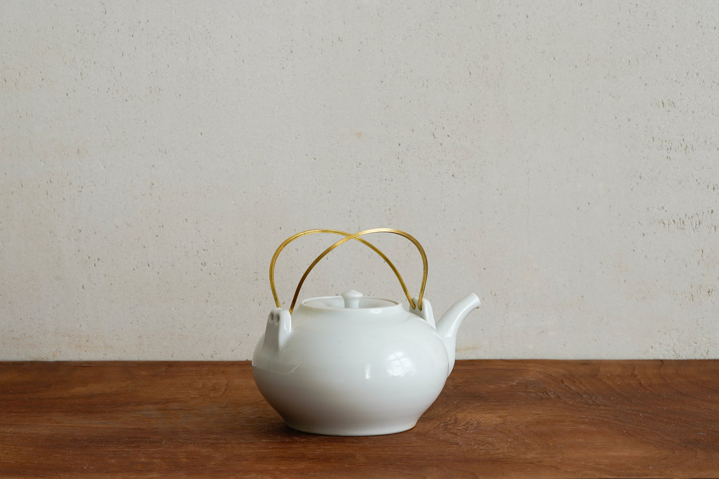 White teapot with gold handle on a wooden surface and beige wall background, from Yuichi murakami